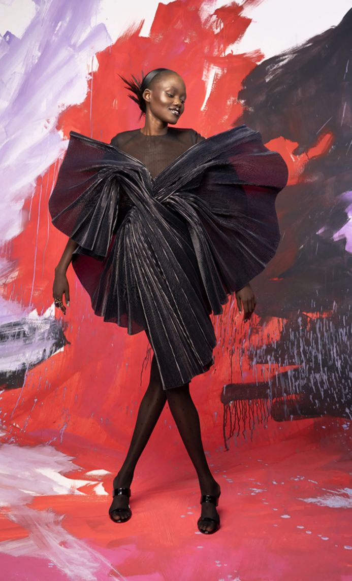 a black woman in a black pleated dress stands in a studio with an abstract background. the woman poses with one hand on her hip and the other by her side. the background is a mix of red, purple, and white. the dress is very  dramatic and flouncy and the model is looking at the camera with a serious expression. she is wearing black heels. the image is a fashion photograph and it is very stylish and sophisticated.