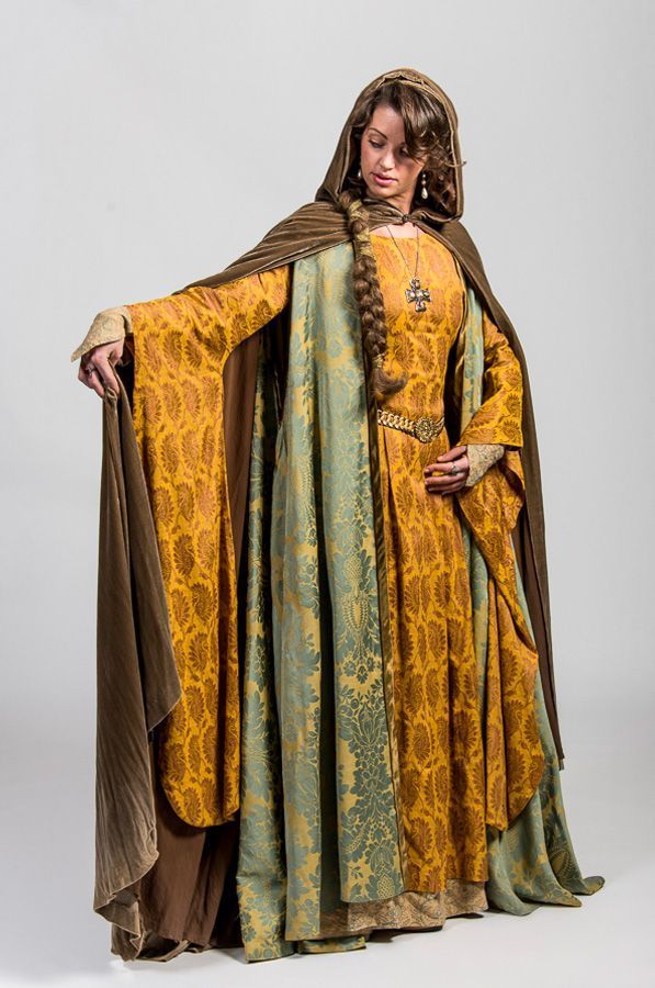 a woman is wearing a medievalstyle dress with a brown hooded cloak. the dress is gold with a brown damask pattern, and has wide, flowing sleeves with lace at the wrists. a decorative belt cinches the waist. a green and gold damask panel is visible down the center of the dress. she wears a long braid and a cross necklace. the background is a plain white.