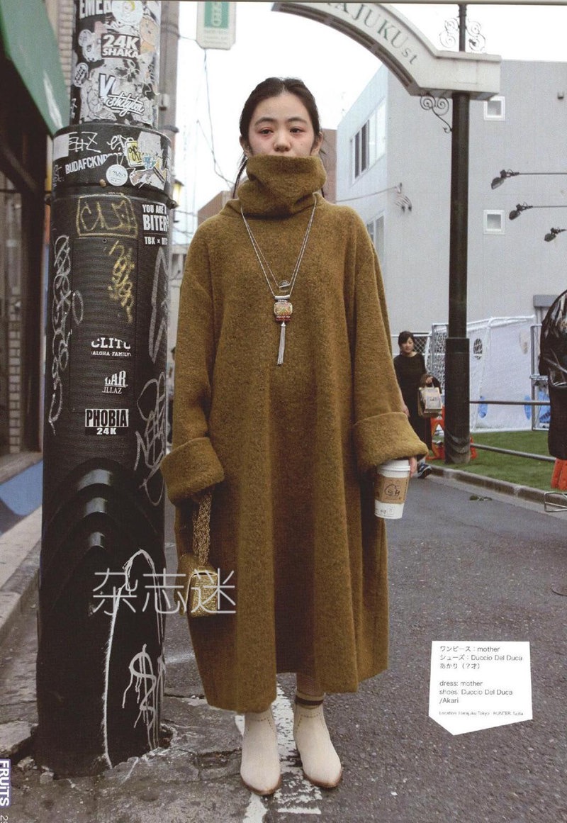 a young woman poses on a city street, showcasing a stylish winter outfit. she wears a long, oversized brown turtleneck dress that reaches her ankles. the dress features dropped shoulders and long sleeves with ribbed cuffs. she pairs the dress with white ankle boots for a chic contrast. her accessories include a long pendant necklace and a woven handbag. her hair is styled simply, and her makeup is minimal. the background features a bustling city street with japanese signage and graffiti adding to the urban ambiance.