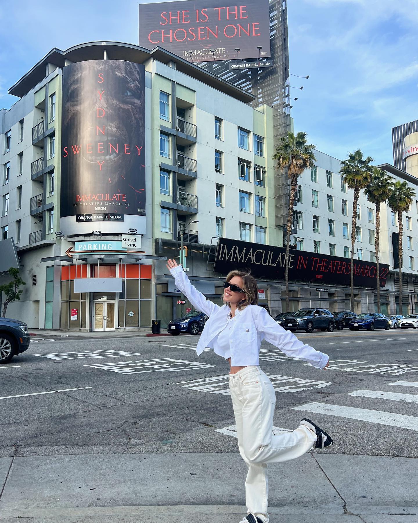 the image shows a young woman joyfully posing in the middle of a los angeles street, with large billboards advertising the movie "immaculate" featuring sidney sweeney.  the woman is wearing a white cropped shirt and loosefitting offwhite pants.  she's smiling, arms outstretched, and appears carefree.  the background is dominated by a multistory building displaying both a large image and smaller text related to the film, along with other advertisements. the scene is a typical city street, with parked cars and crosswalks visible. the overall atmosphere is bright and cheerful, emphasizing the movie promotion.