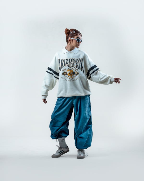 a young woman is posing in a studio against a white background. she is wearing a white crewneck sweatshirt with black stripes and an arizona phoenix graphic. the sweatshirt is oversized, and she is pairing it with loosefitting teal pants and grey adidas sneakers. she's wearing sunglasses and has her hair in a bun. the overall style is casual, sporty and slightly retro, with a vintage 90s vibe. the image is welllit and showcases the model's outfit and pose effectively.