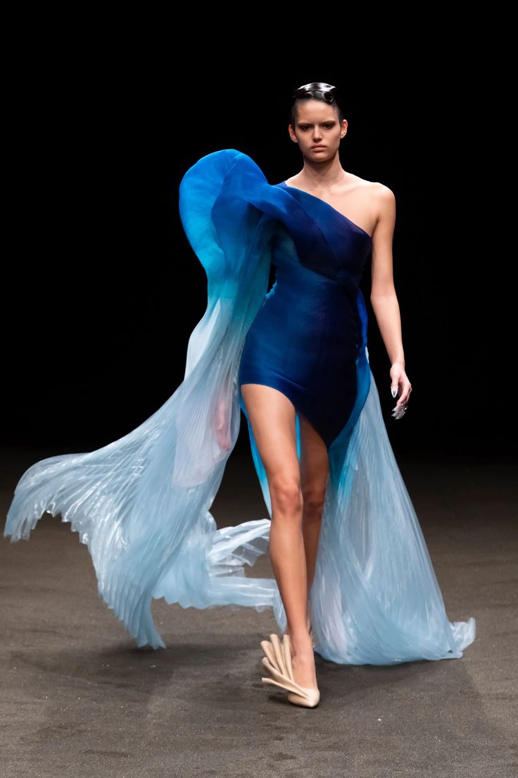 a woman in a blue dress and high heels is walking the runway. the dress is a oneshoulder design with a blue gradient and a flowing train. the woman is posing for the cameras and looking confident. the dress is designed to be very eyecatching and flowy. the woman's hair is black and sleek, and her makeup is simple and elegant. the dress is a beautiful example of high fashion. the dress is designed to be very eyecatching and flowy. the woman's hair is black and sleek, and her makeup is simple and elegant. the dress is a beautiful example of high fashion. the dress is designed to be very eyecatching and flowy. the woman's hair is black and sleek, and her makeup is simple and elegant. the dress is a beautiful example of high fashion.