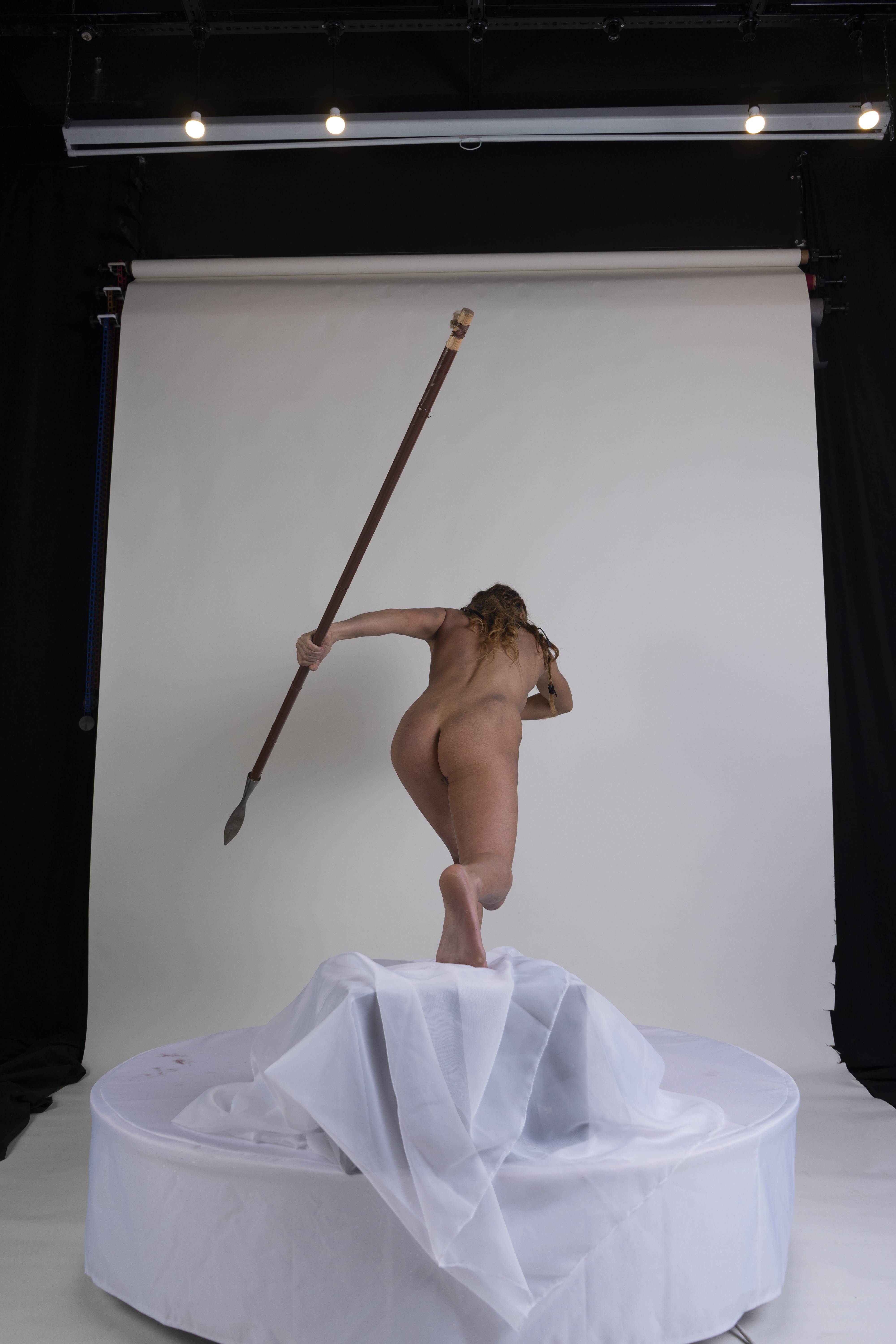 a nude woman stands on a white platform covered in flowing white fabric, holding a spear. she is seen from the back in a studio setting with a white backdrop. the lighting is dramatic, casting shadows and highlighting her form.