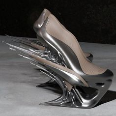 a pair of silver high heels with an abstract design. the shoes are made of a metallic material and have a sharp, pointed toe. the heels are very high and thin, and they extend up the back of the foot in a series of curves and spikes. the shoes are shown on a plain, lightcolored surface.