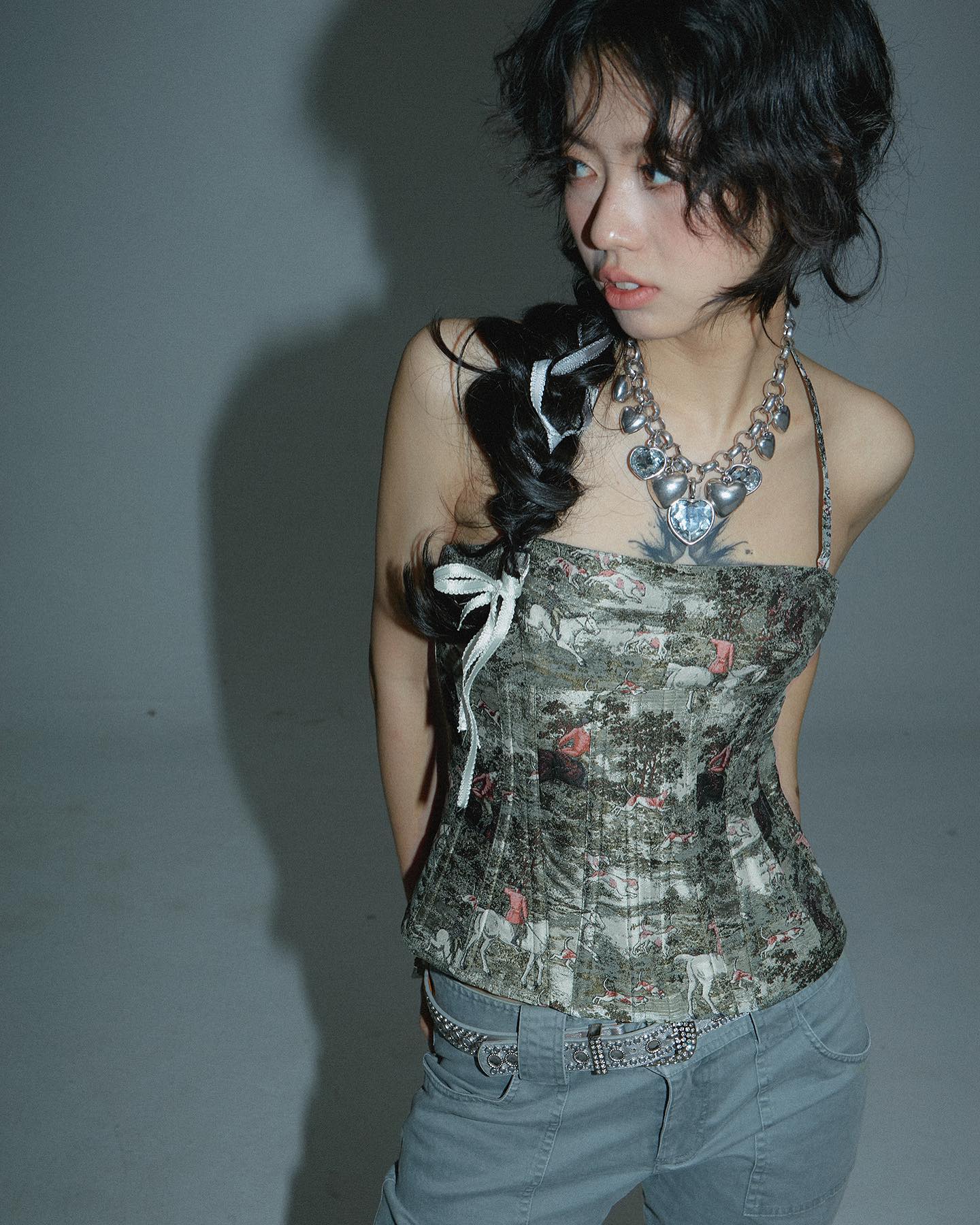 young woman with dark hair styled in a braid is shown from the waist up against a grey background.  she's wearing a green and grey floral print corset top with a silver belt. she also has on grey jeans and a silver heartshaped necklace. her expression is serious and she is looking off to the side.  the overall style is edgy and fashionable.  she has a small tattoo visible on her chest.