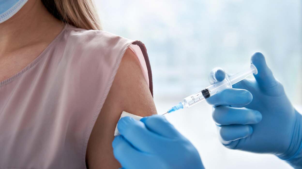a closeup shot shows a person receiving a vaccine. a medical professional wearing blue gloves holds a syringe and is injecting it into the patient's upper arm, which has been cleaned with a cotton swab.