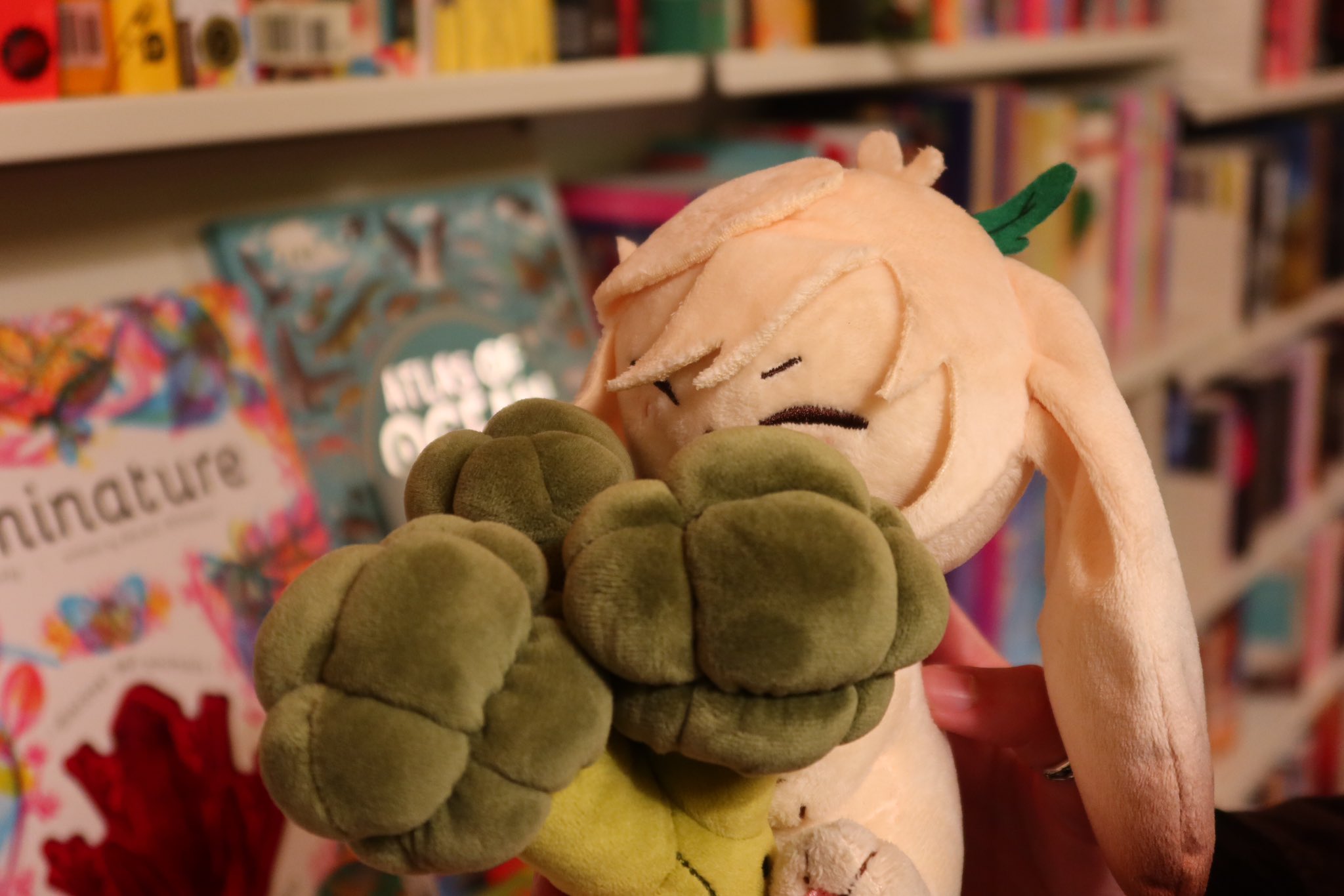 a white plush bunny toy with green broccoli ears, is sitting on a bookshelf full of colorful books. the bunny is cuddly and soft, and its face is slightly tilted with its eyes closed, creating a relaxed and adorable expression. the bookshelf, filled with various books of different colors and sizes, serves as a vibrant backdrop. the overall scene conveys a sense of warmth and comfort, highlighting the allure of soft toys and the charm of a wellstocked bookshelf.