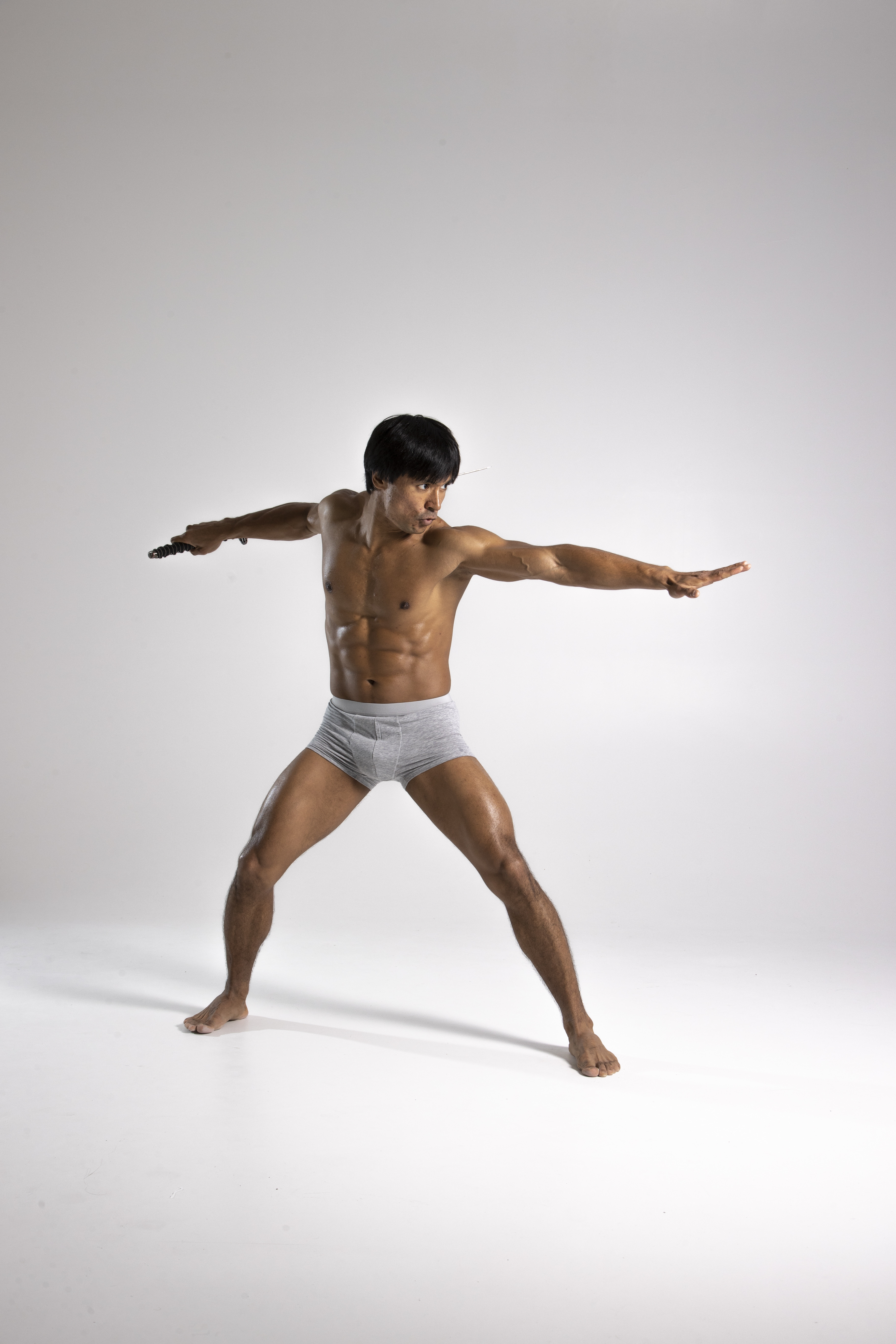 a muscular man is in a studio shot, isolated on a white background. he is barechested, wearing gray underwear, and holding nunchucks. he is in an action pose, with his arms extended and legs spread, showcasing his athletic build and toned physique. the man has dark hair and appears to be engaged in a martial arts practice.