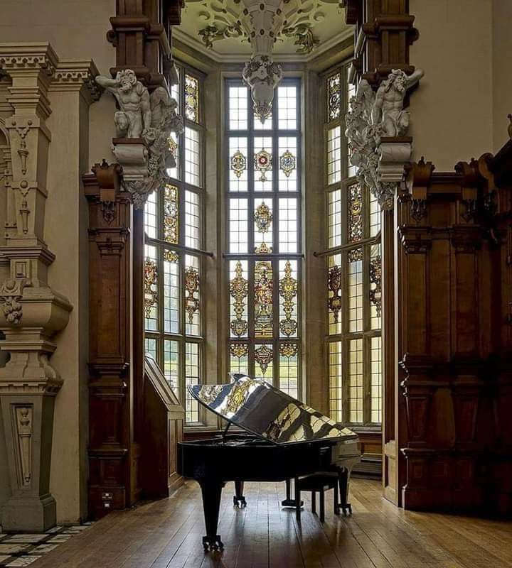 the image showcases a large, polished grand piano positioned within a magnificent room featuring a stunning bay window with intricate stained glass.  the room itself boasts highly detailed, dark wood paneling, elaborate carvings, and sculpted figures adorning the architectural elements. the overall aesthetic is one of classical grandeur and opulence, suggesting a historical setting like a palace or stately home. the contrast between the reflective piano and the warm tones of the wood, coupled with the colorful stained glass, creates a visually captivating scene.  the natural light streaming through the window illuminates the room, highlighting the intricate details and craftsmanship.