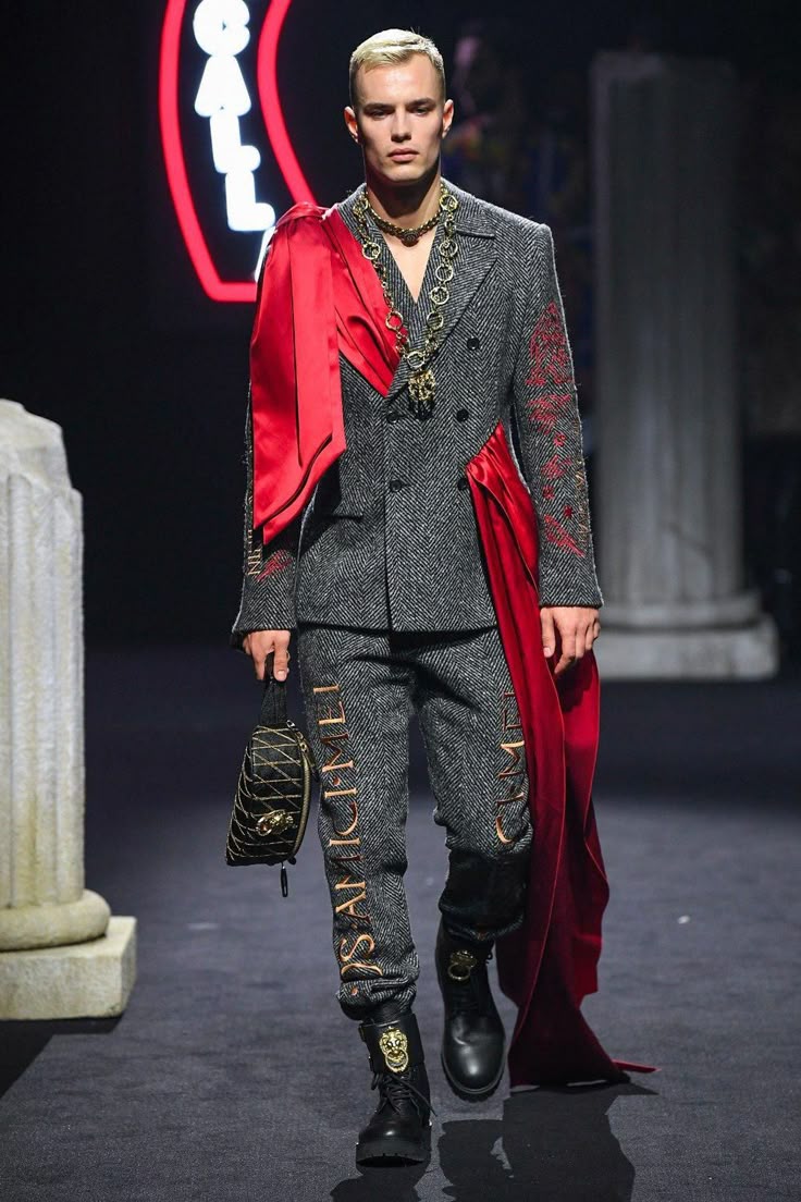 a male model walks down a runway wearing a gray tweed suit with gold lettering on the pants, a red sash draped over one shoulder, gold chains, black boots, and a black handbag.