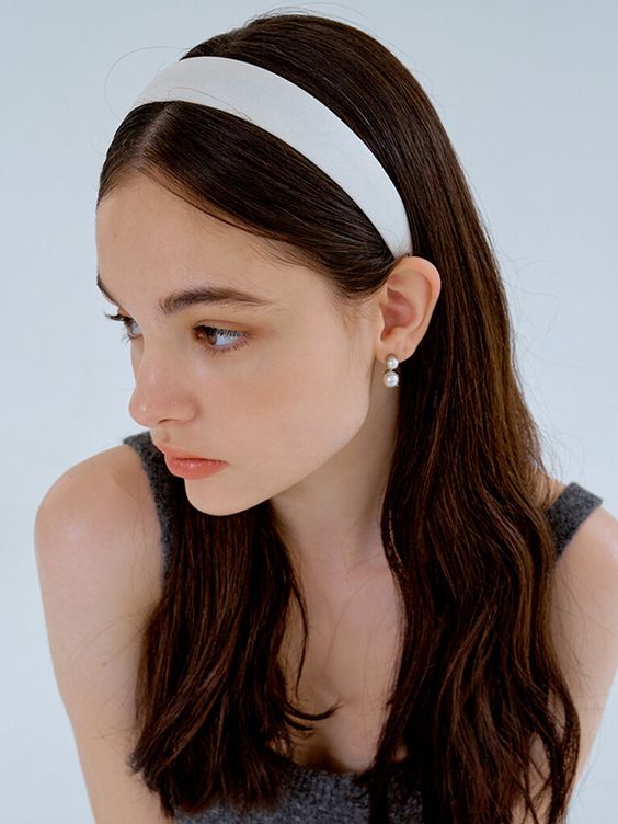 a young woman with long, dark brown hair is wearing a white satin headband. she is wearing a simple pearl earring and a gray tank top. she has a neutral expression on her face and is looking to the side. the background is white and the lighting is soft and natural.