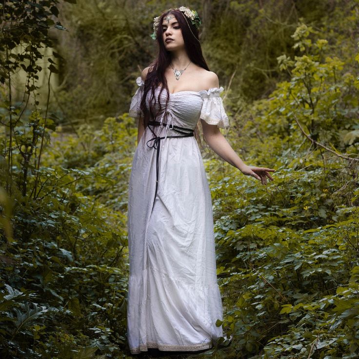 a young woman with long dark hair and a flower crown stands in a lush green forest, wearing a flowing offtheshoulder white maxi dress. the dress has a cinched waist with a dark belt and a slightly gathered bodice. she's adorned with a delicate silver necklace.  the overall scene evokes a fairytale or mystical atmosphere, with the woman appearing ethereal and elegant amidst the natural surroundings.  the lighting is soft and diffused, creating a dreamy, romantic mood.