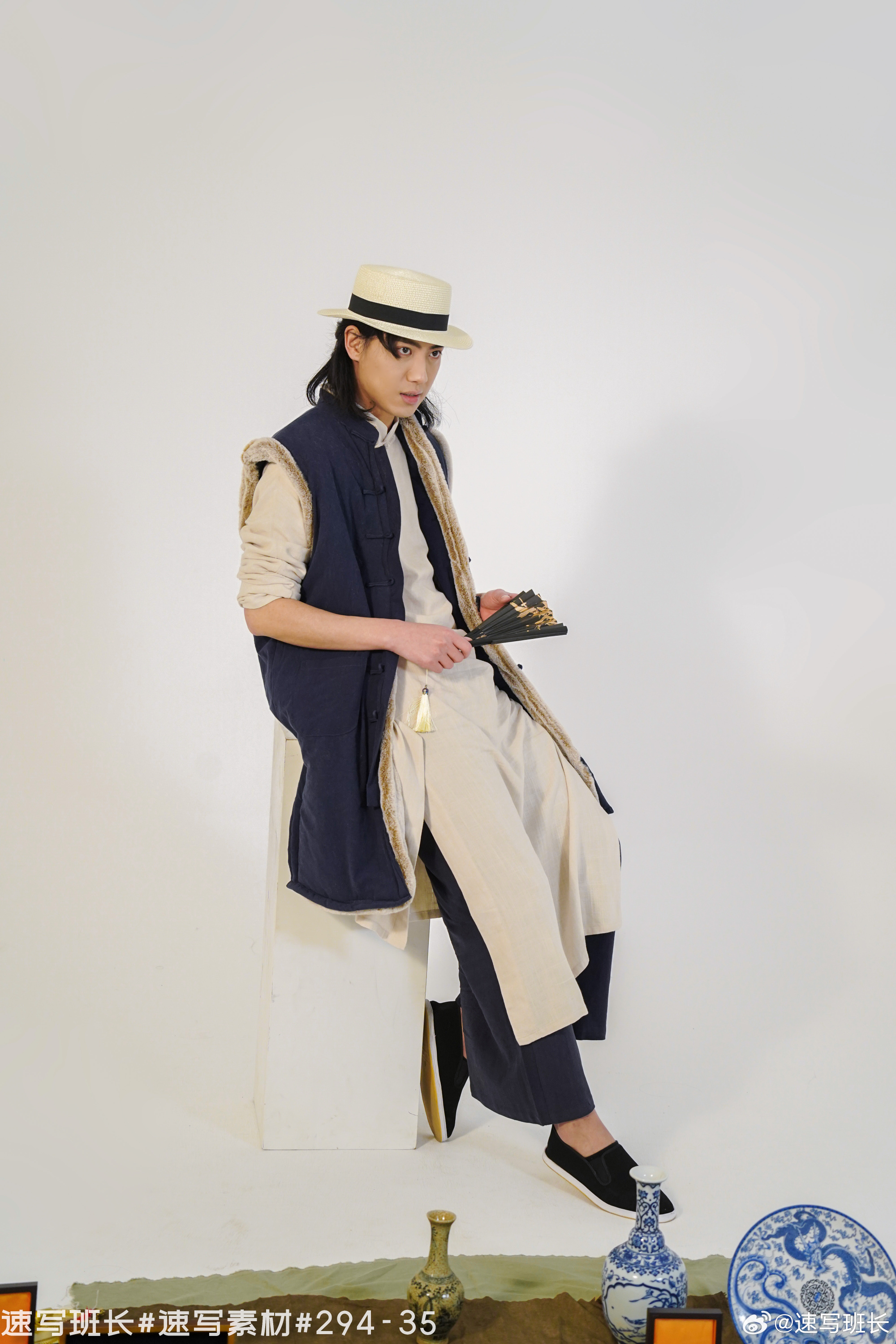 a man wearing a straw hat, a dark blue vest with fur trim, a long white robe, and white shoes is seated on a white cube in a studio setting. he is holding a black and gold fan in his right hand. there are two blue and white porcelain vases and a blue and white porcelain plate on the floor in front of him. the background is white.