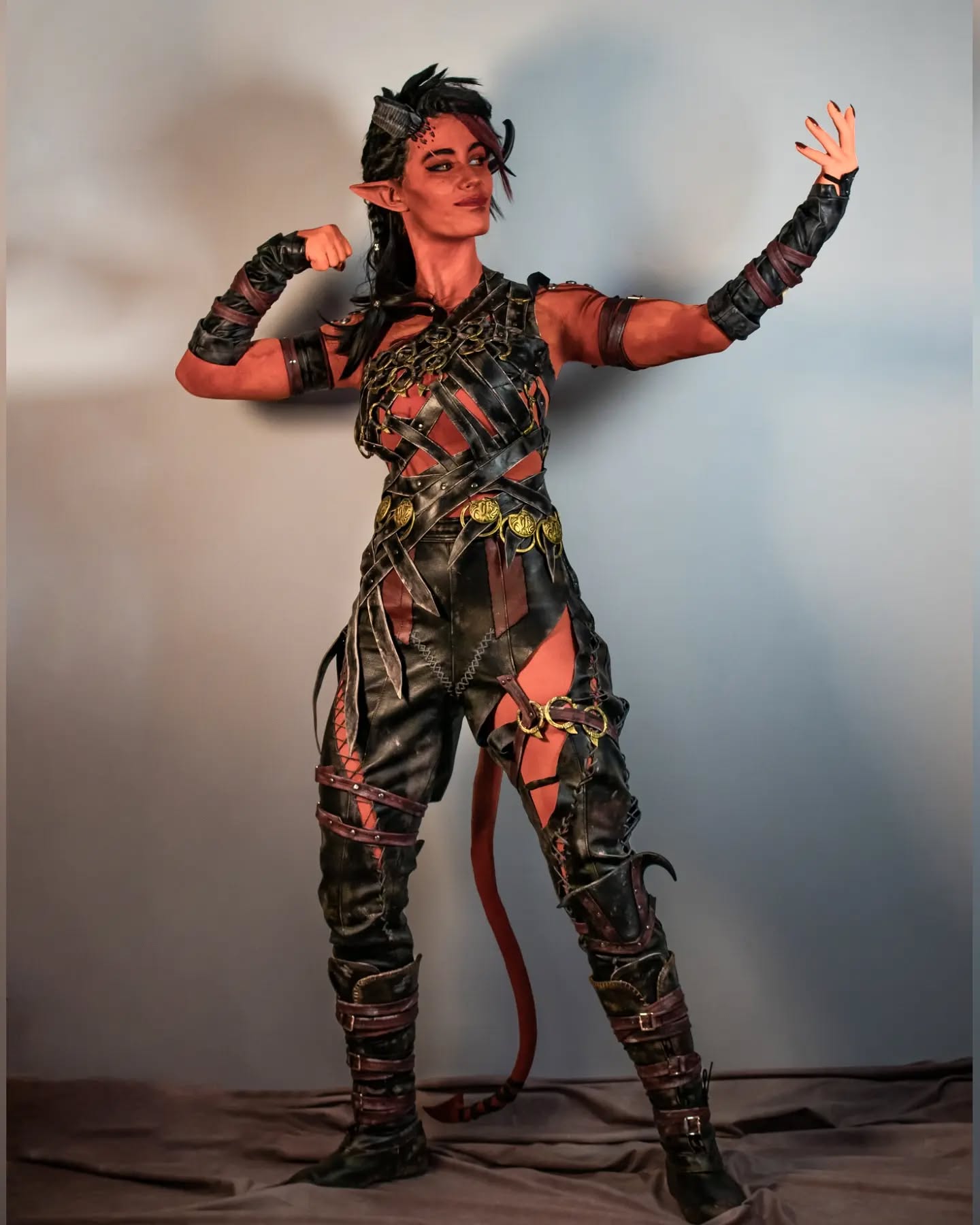 a fullbody shot of a female cosplayer portraying a tiefling character. the cosplayer has red skin, horns, and a tail, and is wearing a meticulously crafted leather armor costume with intricate details.  the armor is dark in color with gold accents. the pose is dynamic, suggesting strength and power. the background is a simple, light gray studio backdrop. the overall image conveys a sense of fantasy and immersive roleplaying.