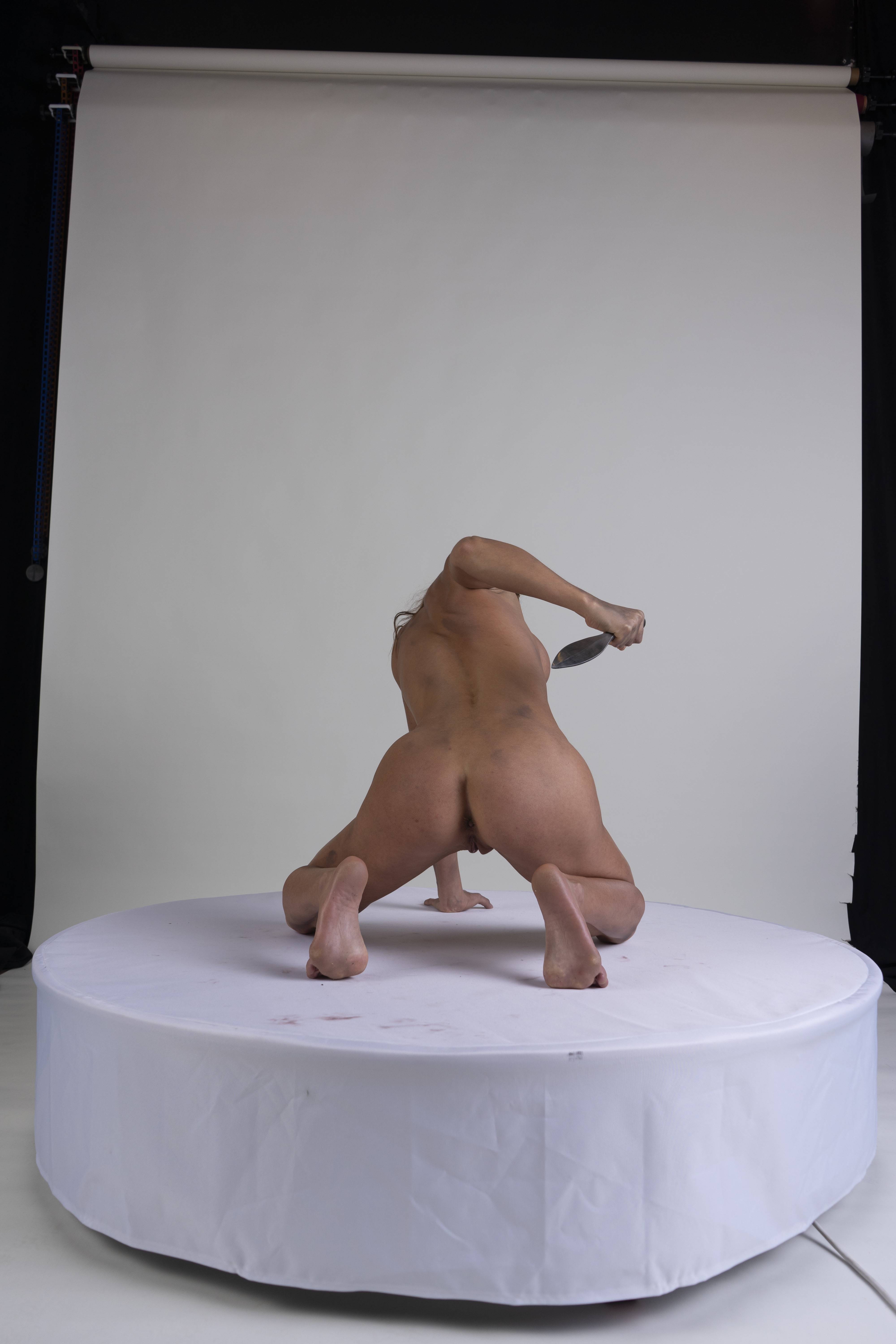 a nude woman is kneeling on a white circular platform with her back to the camera. she is holding a knife behind her back. the background is a white studio backdrop.