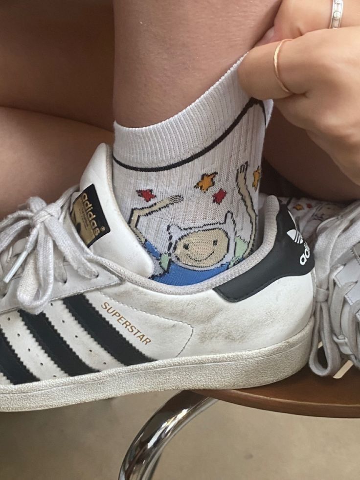 a closeup shot shows a person wearing white socks with a cartoon graphic, paired with white adidas superstar sneakers. the person is holding the top of the sock, which features finn the human from adventure time and a few red and yellow maple leaves. the sneakers have the classic black stripes and black heel tab, along with the "superstar" text on the side. a couple of rings are visible on the person's fingers. the footwear is placed on a wooden surface with a chromecolored leg of a chair visible underneath.