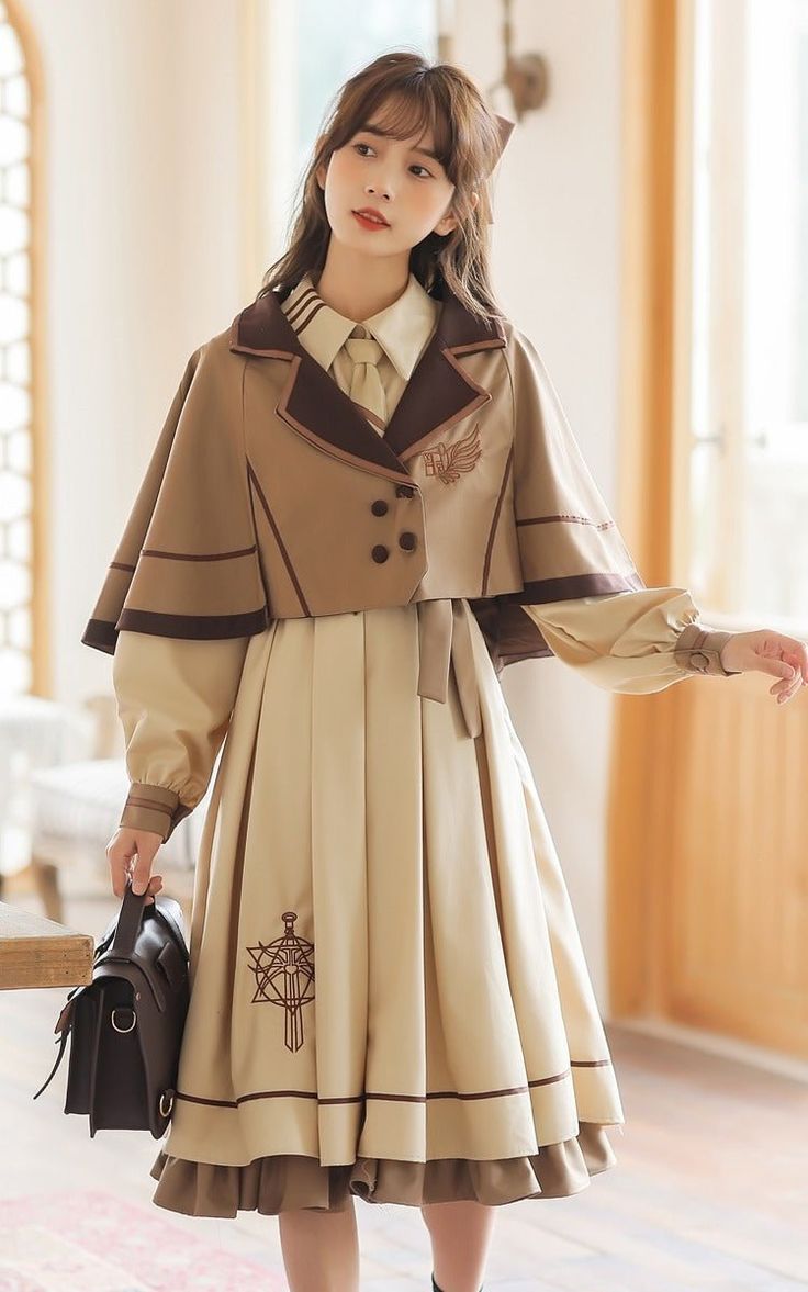 a young woman is posing in a vintageinspired outfit, typical of lolita fashion. she is wearing a beige dress with brown accents, a matching cape, and a white shirt with a brown collar. the dress and cape feature embroidered details, including swords and wings. she is also carrying a brown leather purse. the setting appears to be indoors with studio lighting.