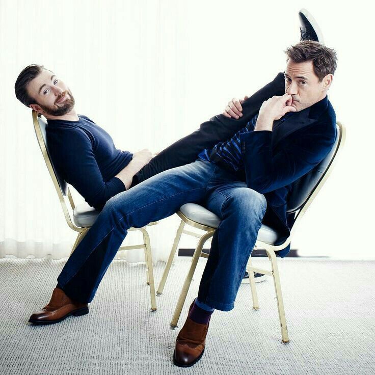 chris evans and robert downey jr., known for their roles as captain america and iron man respectively, are seen sitting on chairs during a photoshoot. evans is reclining on a chair while downey jr. is sitting upright, holding evans' legs. both actors are dressed in casual attire. they are both smiling, and evans is looking at the camera.
