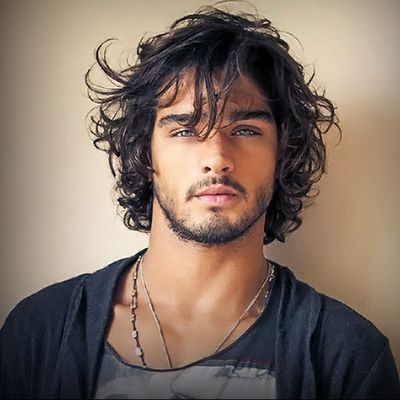 a portrait of a man with wavy dark hair, blue eyes, and a beard and stubble. he is wearing a casual black shirt and two necklaces. the background is a neutral beige color.