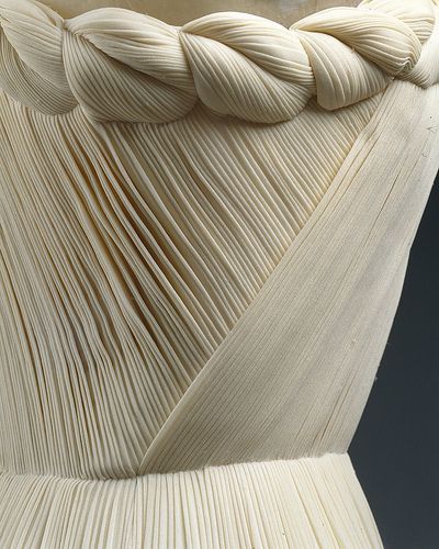 a closeup image shows the bodice of a creamcolored dress. the fabric is heavily pleated, creating a linear texture. a draped sash or band crosses the bodice diagonally. the top of the dress features a decorative rope detail, resembling a braid or twisted cord.