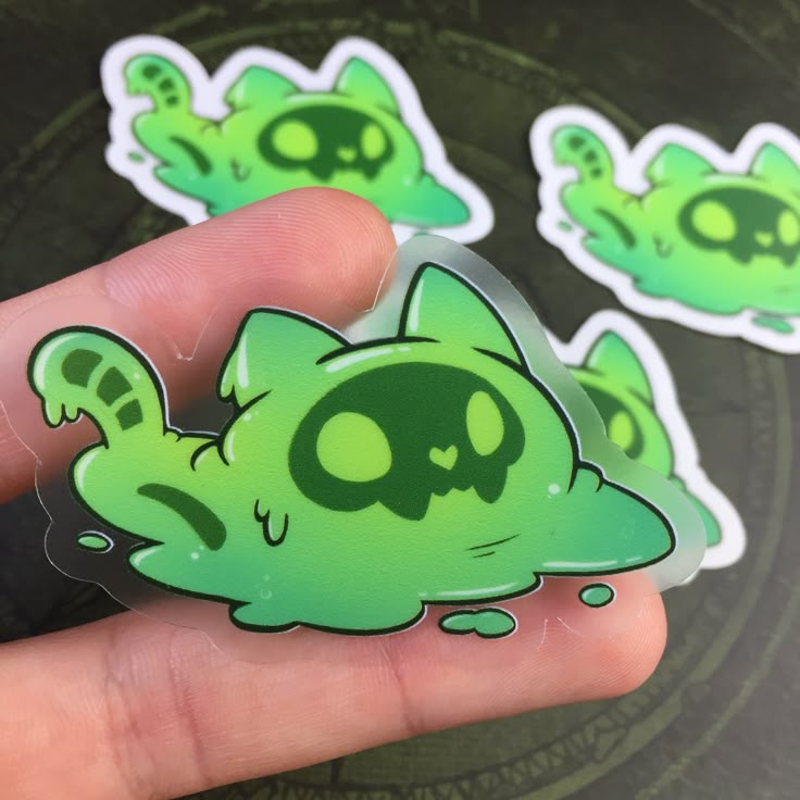 the image shows stickers of a cat made of green slime. the cat's face is a skull shape with heartshaped nose and round eyes. the stickers have a transparent background and a white border. a person's hand is holding a slime cat sticker.
