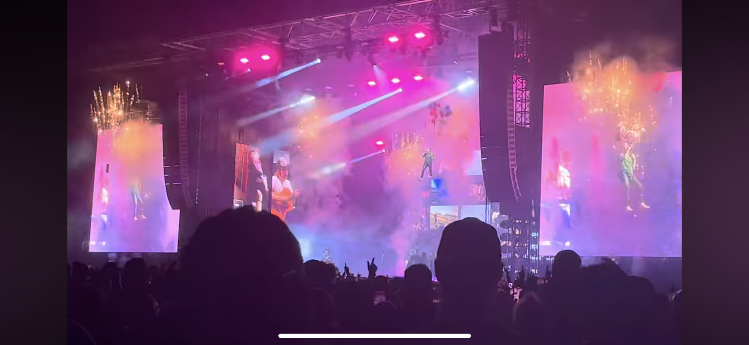 this is a photo taken at a concert. the photo captures a singer on a screen, with a crowd of people in front of the stage, facing the stage. the stage is lit with pink and purple lights, and there is smoke in the air. the photo is taken from the perspective of someone in the audience.