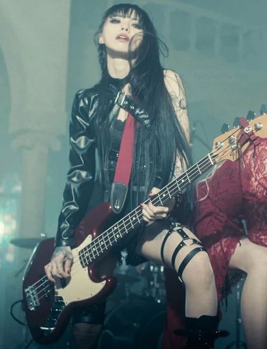 a woman with dark hair and tattoos is playing a bass guitar on stage. she is wearing a leather jacket, a short black dress, garter and boots. another woman in a red dress is next to her. there are drums and other stage equipment in the background.