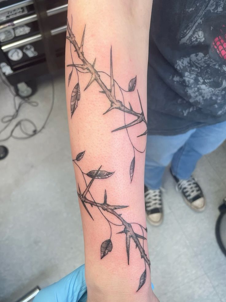 a closeup shot of a person's left forearm tattoo, showcasing a black and grey intricate design of a vine with thorns and leaves, signifying a modern and minimalist aesthetic. the image captures the delicate details of the tattoo art, showcasing the artist's skill and creativity. the tattoo extends down the forearm, highlighting the beauty of the art on the human body.