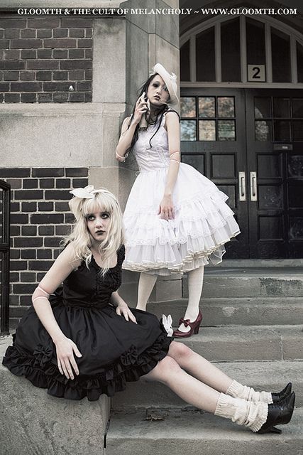 two girls in lolita fashion stand in front of a brick building. the girl on the left wears a black dress and is sitting on the stairs. the girl on the right wears a white dress and is standing with one foot on the stairs. both girls have long hair. the background is a brick wall and a door.