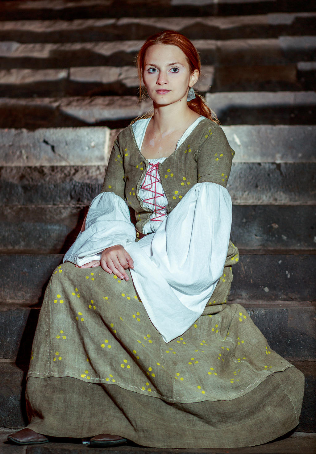 a woman in a historical dress sits on stone stairs. she has red hair and is wearing a long, olive green gown with yellow accents and a white undershirt with red lacing. the setting appears to be a castle or old building.