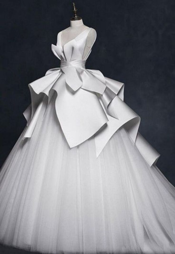 a stunning white bridal gown with a dramatic, architectural design. the bodice is made of smooth satin and features a single, draped strap. the skirt is a layered tulle with a dramatic satin ruffle that creates a flowing, dramatic effect. the model is standing in a studio, with a black background, showcasing the dress's elegance and beauty.