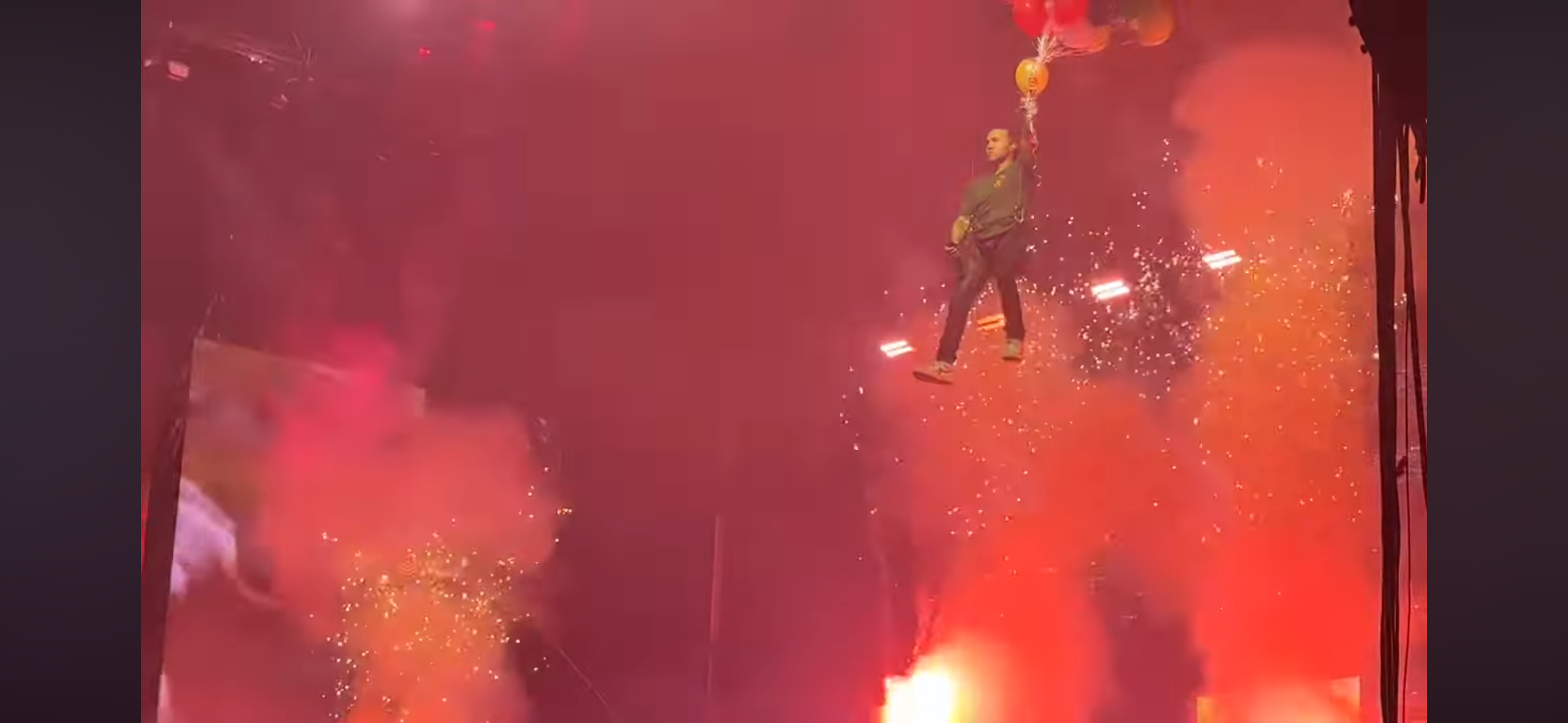 a man is suspended in the air above a concert stage with multiple balloons attached to him. there is a dense smoke cloud beneath him, and fireworks and confetti rain down on him. the stage is brightly lit and surrounded by a large, excited audience.