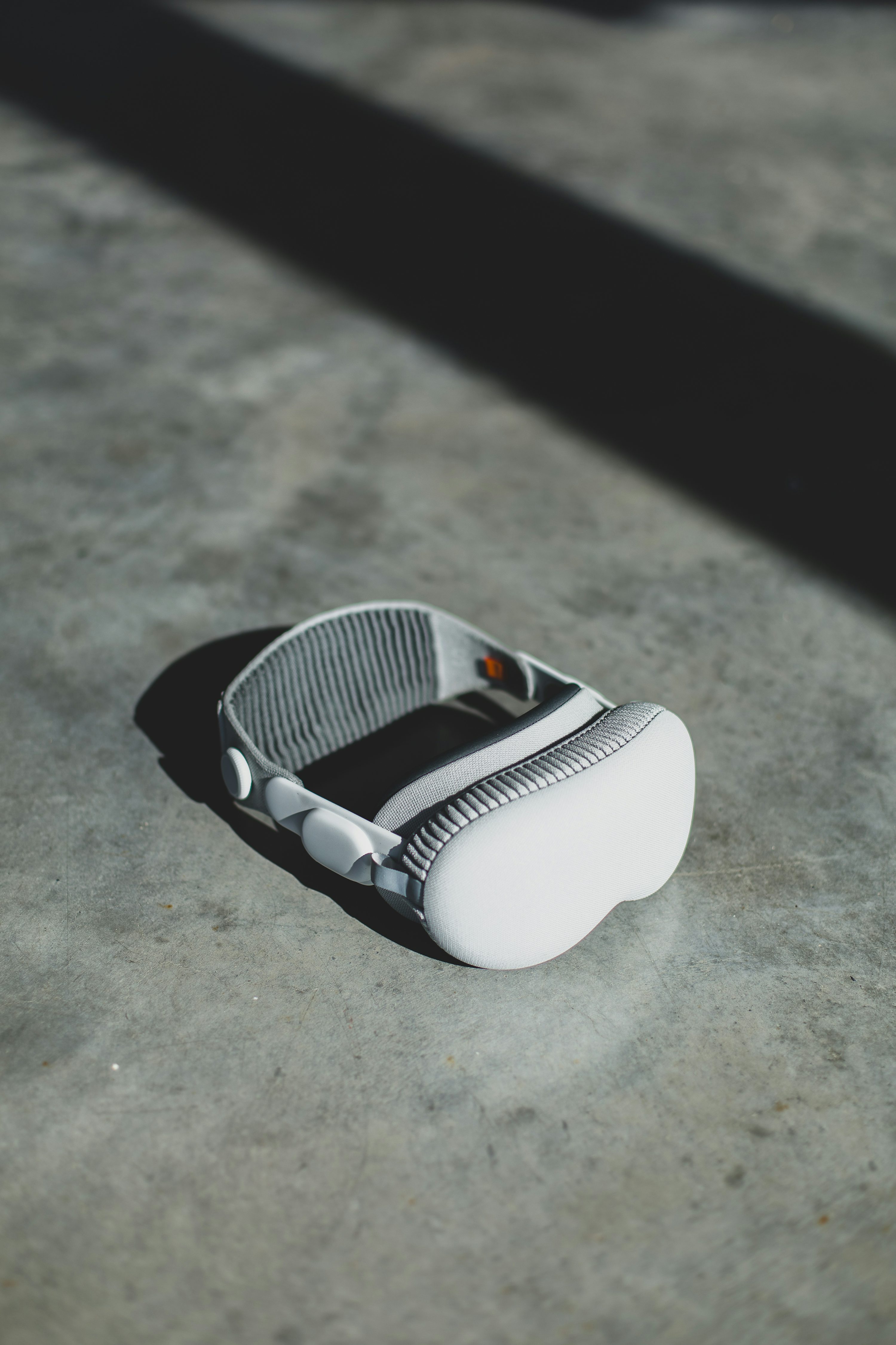 the image shows an apple vision pro vr headset resting on a gray, textured surface. a shadow stretches across the surface, partially obscuring the headset. the headset is white and gray with a fabriclined headband.