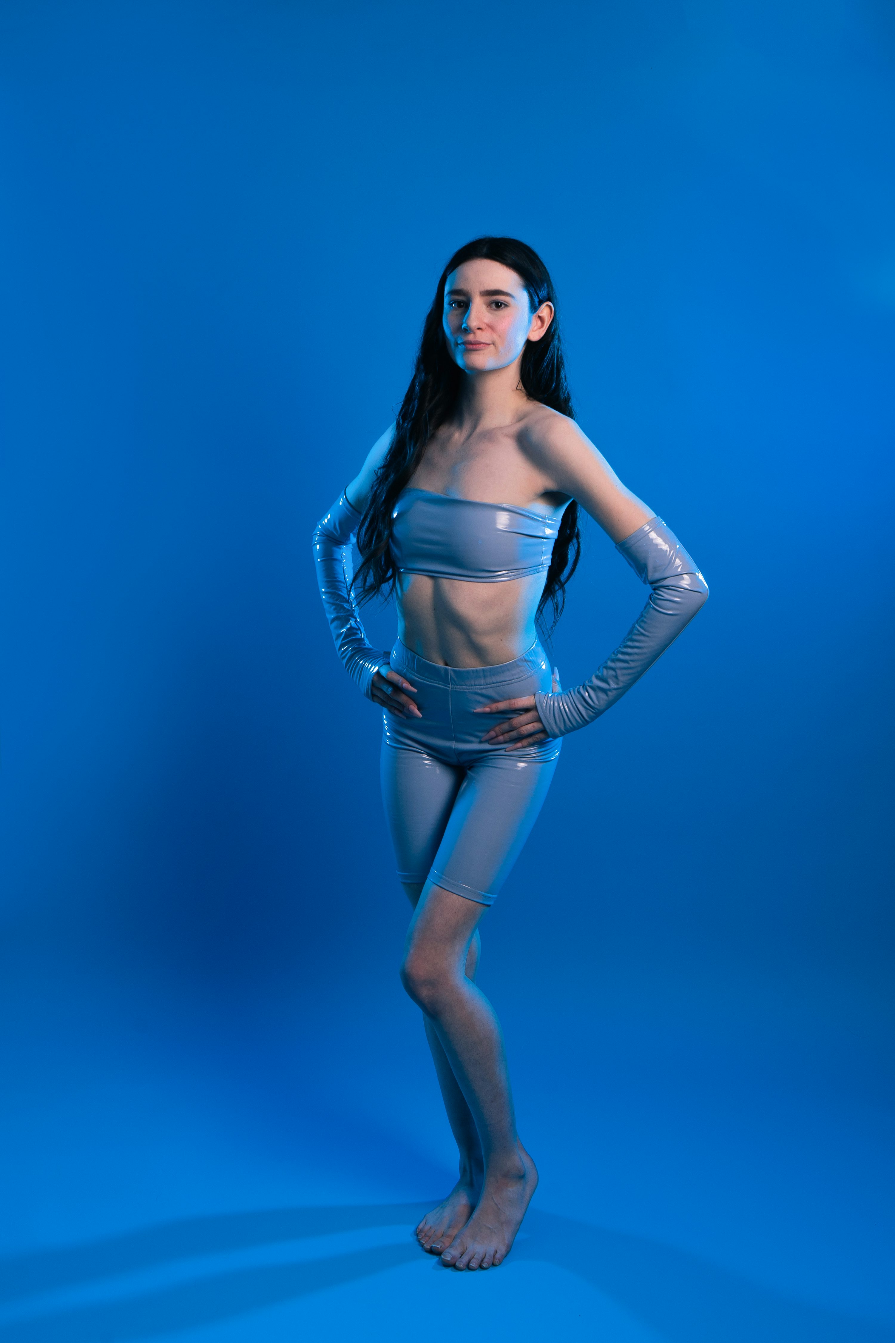 a full shot of a young woman with long dark hair posing against a bright blue background. she's wearing a formfitting light blue outfit that looks like it's made of a shiny material possibly latex or vinyl. the outfit includes a strapless top and highwaisted shorts. she has long sleeves that go past her elbows. the woman's pose is confident with hands on hips. the image is well lit and the focus is sharply on the model. the overall aesthetic of the image is clean, modern and slightly futuristic.