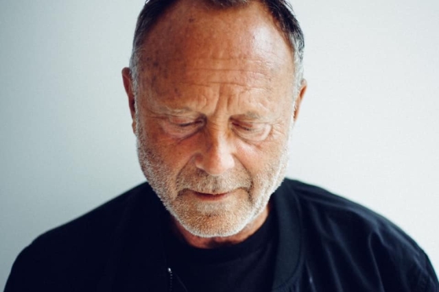 a closeup portrait of an older caucasian man with grey hair and beard. he is wearing a black jacket and is looking down. he has wrinkles around his eyes and on his forehead, and his skin is pale. he appears to be thoughtful or pensive.
