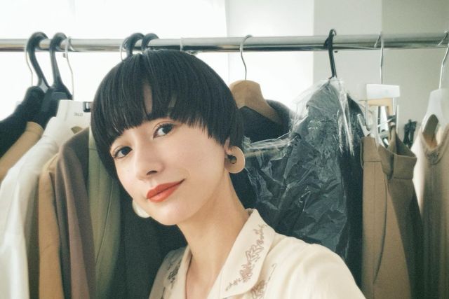 a young woman in a white blouse smiles happily,  while standing in a clothing store with a rack of clothes behind her. the rack of clothes behind her is full of clothing in brown and black. the woman is asian and has short, black hair. she is wearing an oversized white blouse with a floral print.  she is standing in front of a rack of clothes in a clothing store. she is smiling at the camera.