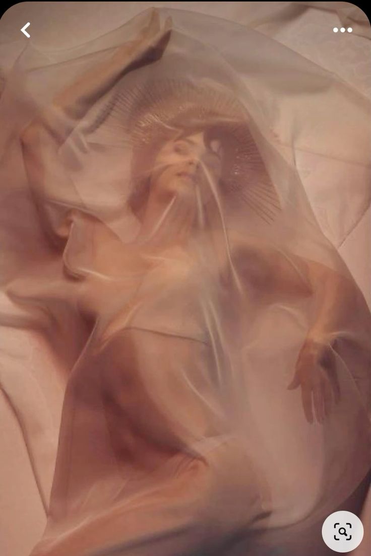 the image is an artistic photograph of a woman seemingly reclining on a bed or surface. she is draped in a flowing, sheer fabric that is translucent, revealing her form beneath but obscuring specific details.  the fabric drapes over her head like a veil or sheet, and she wears a crownlike headpiece. the overall aesthetic is ethereal and dreamlike, using soft, peach and beige tones and subtle lighting to create a mystical and sensual mood. the focus is on the mood and artistic composition rather than sharp detail.