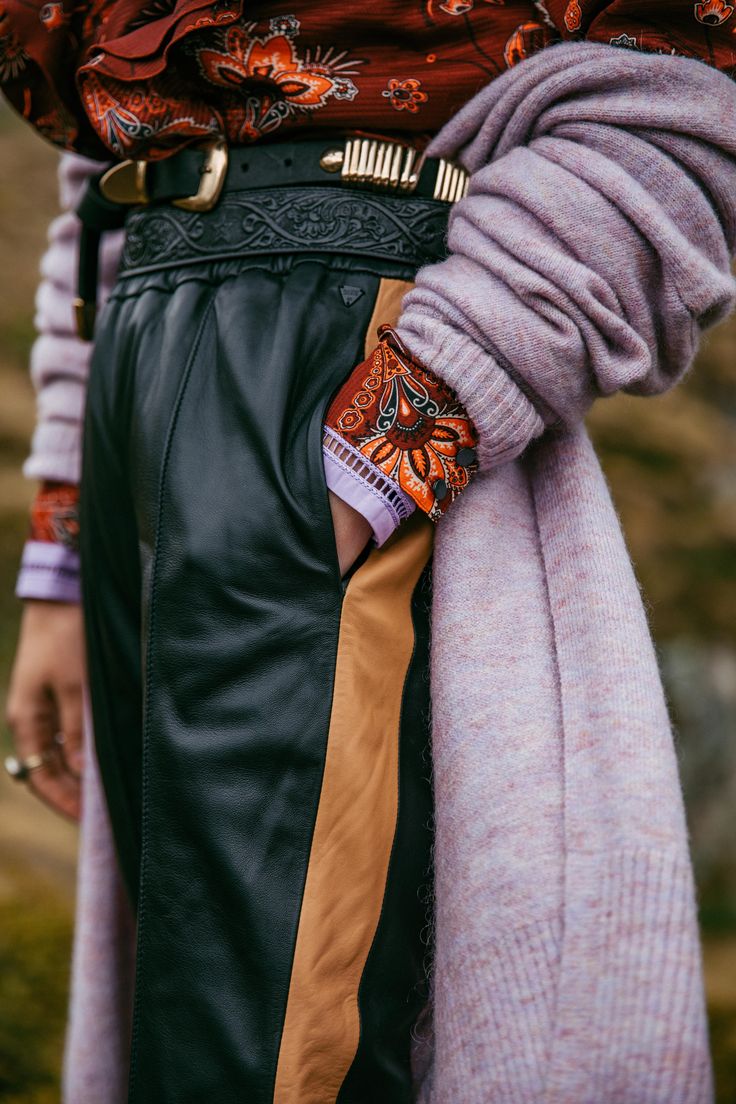 closeup view of a woman's outfit featuring black leather pants with a contrasting tan leather side stripe.  she's wearing an intricately designed black westernstyle belt over a paisleyprinted, longsleeved shirt. a large, pale lilac oversized knit sweater is draped over her shoulders and arms. the image focuses on the textures and details of the clothing, showcasing the rich materials and patterns. the overall style appears to be a blend of western and bohemian influences, suitable for fall or winter.