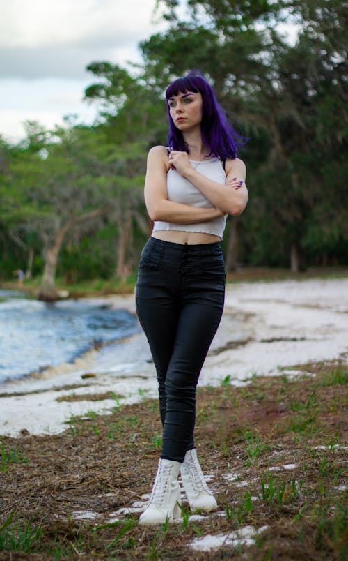 a fullbody shot of a young woman with vibrant purple hair standing outdoors in a natural setting. she's wearing a white crop top, black skinny jeans, and white ankle boots. her arms are crossed across her chest, and she's looking off into the distance with a pensive expression.  the background features trees, a body of water, and grassy ground.  the overall mood is calm and serene.