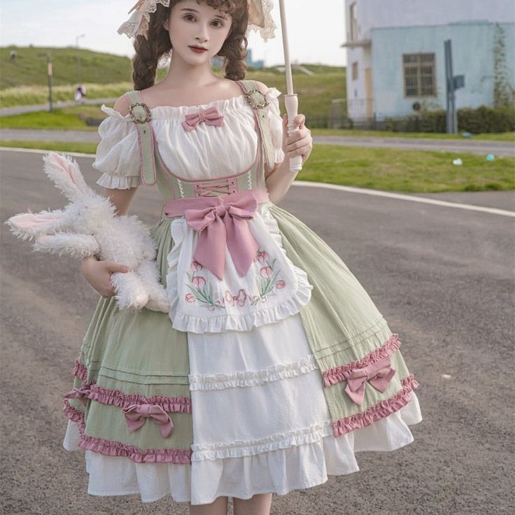 a young woman poses in a green and white lolita fashion dress. she is holding a white stuffed rabbit and a parasol. the dress is adorned with bows, lace, and floral embroidery. she is standing on a paved road with green grass and a white building in the background. the lighting is natural and sunny, giving the photo a bright and cheerful feel.