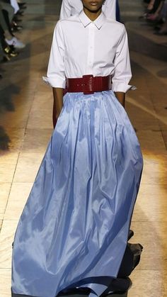 a woman wearing a white longsleeved shirt and a long blue skirt poses for a street photographer during a fashion week. she has a red belt on and is walking confidently, her face turned towards the camera.  the photo captures the beauty of street style fashion and the stylish outfits seen during fashion week.