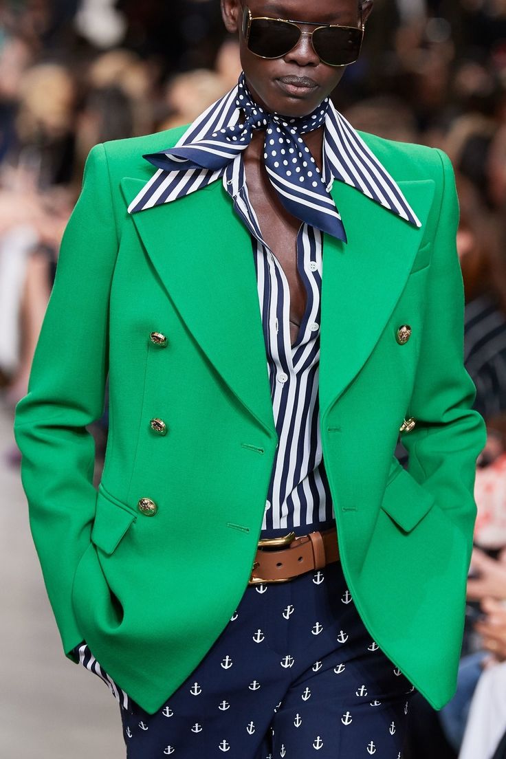 a model is showcasing a vibrant green doublebreasted blazer adorned with gold buttons.  underneath, they are wearing a crisp navy and white striped shirt, accented with a stylish polka dot scarf. the look is completed with navy pants featuring a subtle anchor print and a brown leather belt. the model's sunglasses add a touch of cool sophistication. the overall style is a fresh take on nautical fashion, with bold and vibrant colours. the image appears to be from a fashion show or runway.