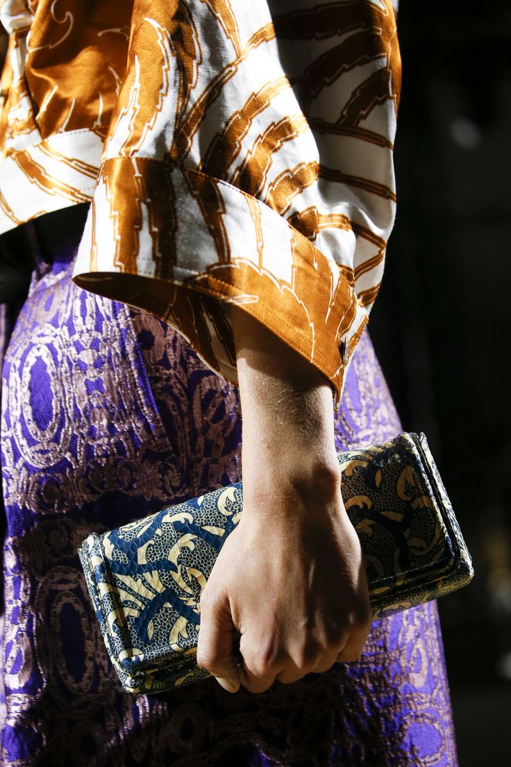 closeup shot of a woman's hand holding a stylish clutch. the woman is wearing a purple patterned skirt and an orange and white patterned top. the clutch is a similar patterned fabric in shades of blue, gold, and offwhite. the image focuses on the textures and patterns of the clothing and accessories. the lighting is dramatic, highlighting the details of the fabrics.