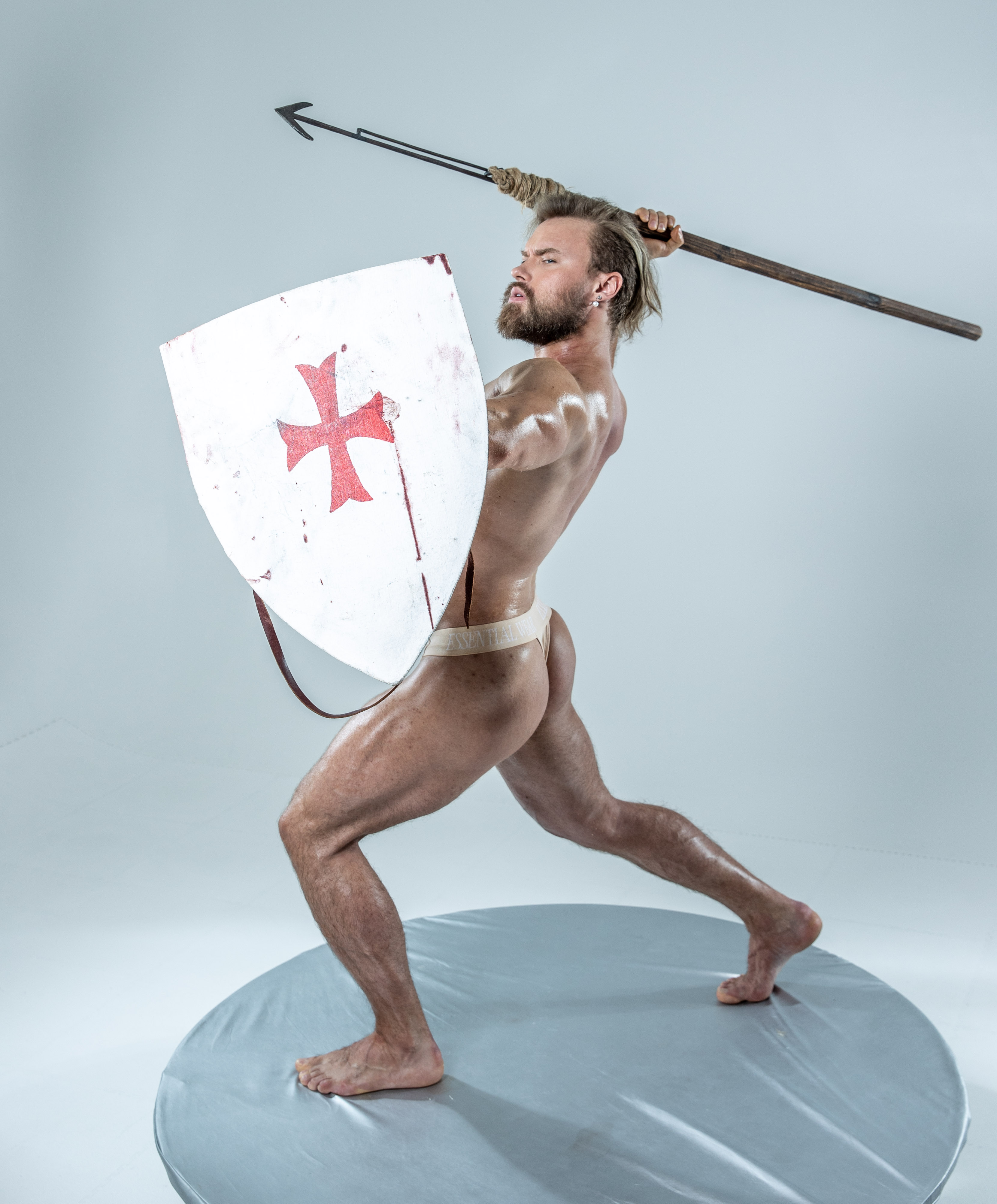 a muscular man, wearing only a loincloth, stands on a grey circular platform against a white background. he is holding a spear in his right hand and a shield with a red cross on it in his left, adopting a dynamic fighting pose.  the image appears to be studiolit, creating a dramatic effect.