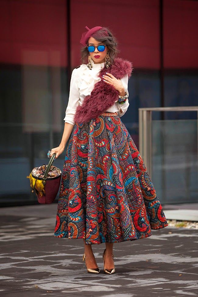a fullbody shot of a stylish woman posing outdoors. she's wearing a white ruffled blouse, a long, colorful paisleypatterned midi skirt, a maroon fur stole, a maroon hat, blue sunglasses, and gold highheeled pumps. she carries a unique, patterned handbag. the background features a modern red and gray building. the overall style is bohemian chic, blending vibrant patterns and textures with elegant accessories. the setting suggests an autumn or winter season. the photo is professionally taken, with good lighting and composition.