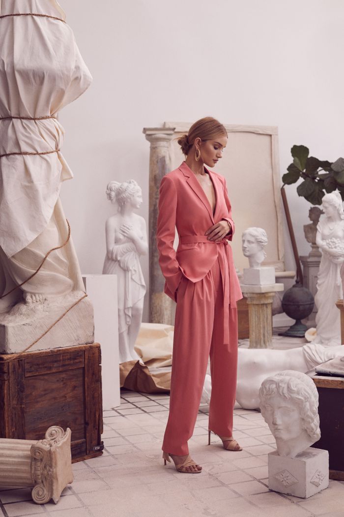 a full shot of a stylish female model wearing a coral pink pantsuit and heels, posing gracefully amongst classical sculptures in a studio setting.  the overall mood is sophisticated and elegant. the model's pose and the surrounding artwork create a visually striking and artistic image.