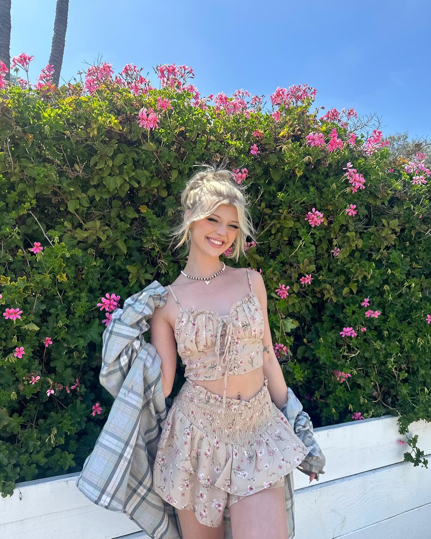 a young woman with blonde hair pulled up in a messy bun smiles in front of a green hedge with pink flowers. she is wearing a floral print crop top and matching shorts. a light plaid buttondown shirt hangs off of her shoulders. she has a delicate necklace around her neck. the sky is blue and sunny. there are trees visible in the background.