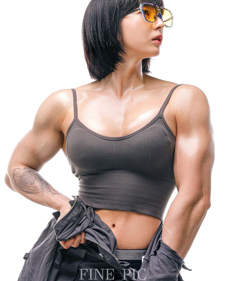 a young woman with short black hair and sunglasses stands with her arms crossed, showing off her muscular arms and abs.  she is wearing a gray tank top and has a strong, athletic physique.  the background is white.