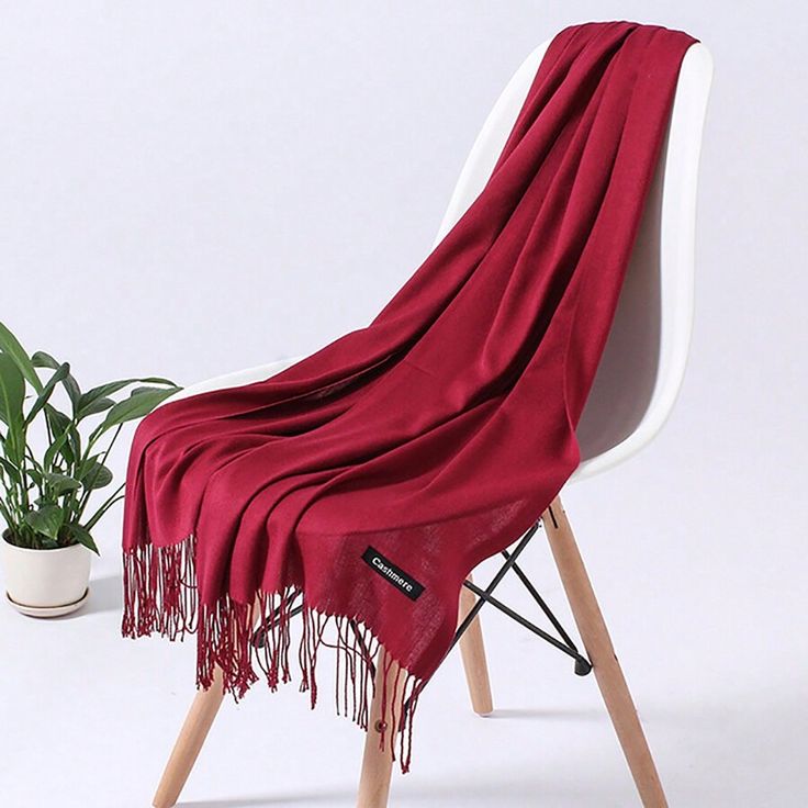 a maroon cashmere scarf with fringe is draped over a white chair. a plant is in a white pot in the background. a small tag is on the scarf with the word "cashmere" on it.