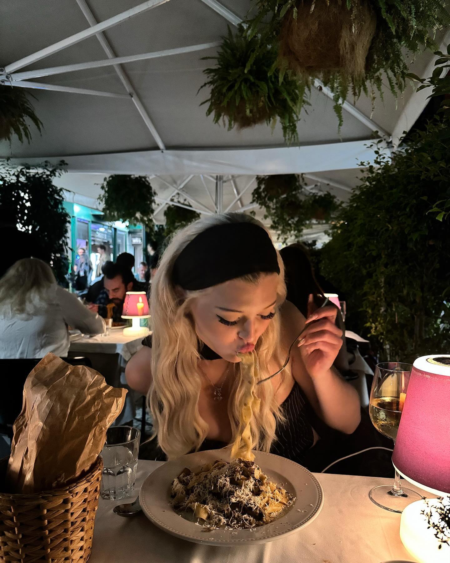 a blonde woman wearing a black headband and choker necklace is eating pasta at a restaurant. she is holding a forkful of pasta up to her mouth. there is a plate of pasta in front of her, along with a glass of wine and a lamp. the restaurant has outdoor seating and is decorated with plants.